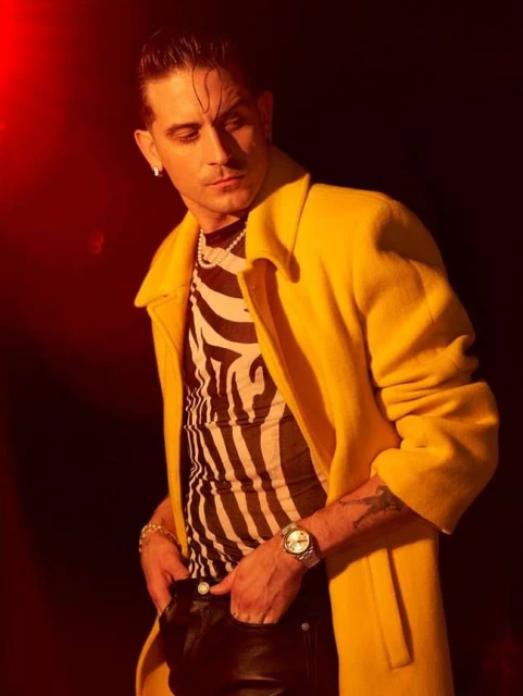 Man in zebra top and yellow coat posing against a dark background with red lighting, wearing jewelry and a watch.