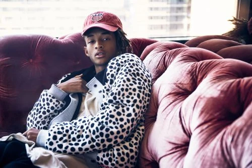 Adult male poses on a pink, tufted couch wearing a red baseball cap and a dalmatian-spotted jacket in an indoor setting with natural light.