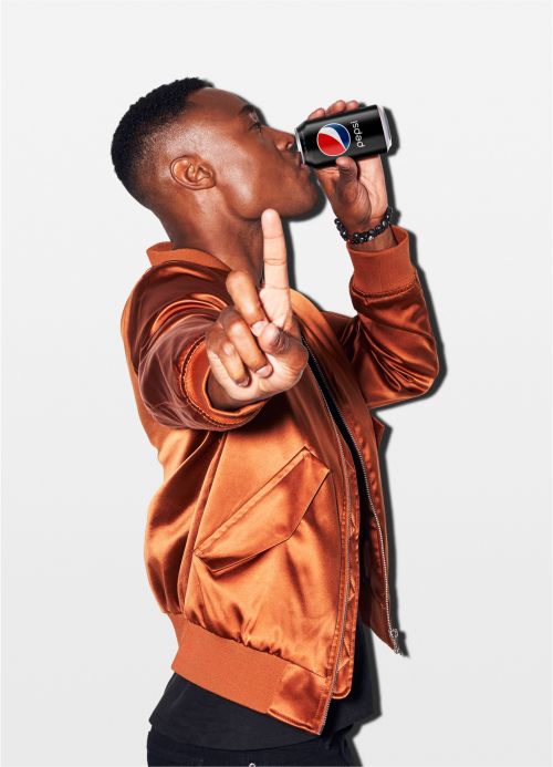 Young man in copper jacket drinks Pepsi can while flashing peace sign in a studio shot with a bright white backdrop.