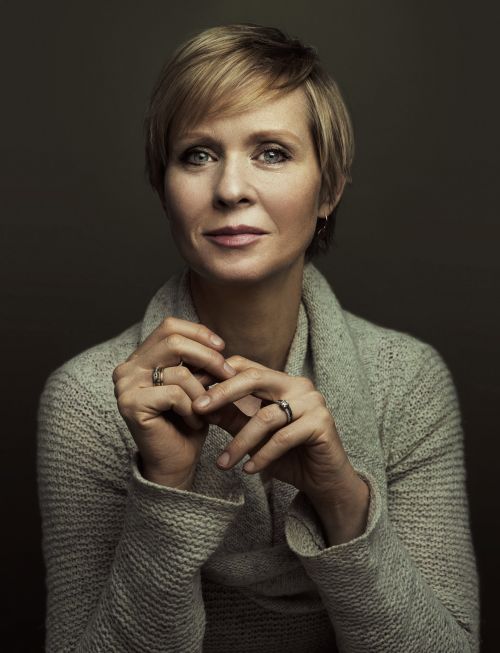Portrait of Jane Lynch in a grey sweater with her hands together, set against a dark grey background in a photo studio setting.