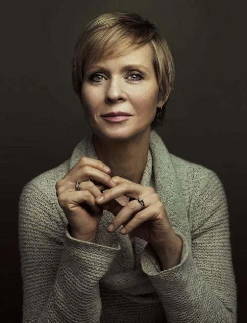 Portrait of Jane Lynch in a grey sweater with her hands together, set against a dark grey background in a photo studio setting.