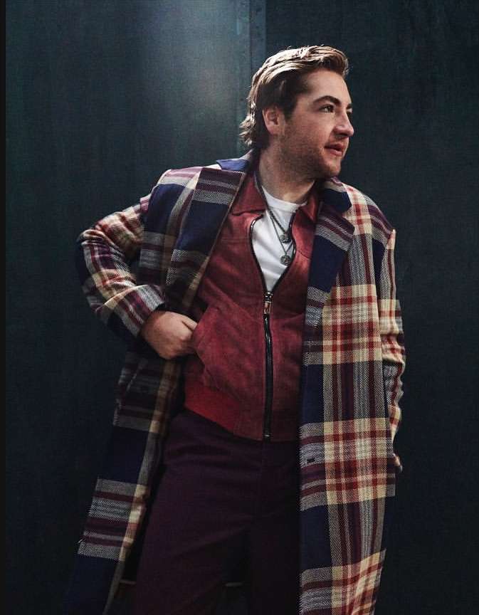 Man wearing a large plaid coat and red vest, looking off to the side with a slight smile, in a studio or indoor setting.