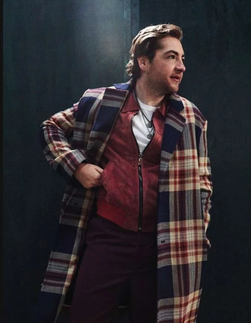 Man wearing a large plaid coat and red vest, looking off to the side with a slight smile, in a studio or indoor setting. 