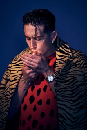 Man lighting a cigarette wearing an animal print coat and a red spotted top against a blue background