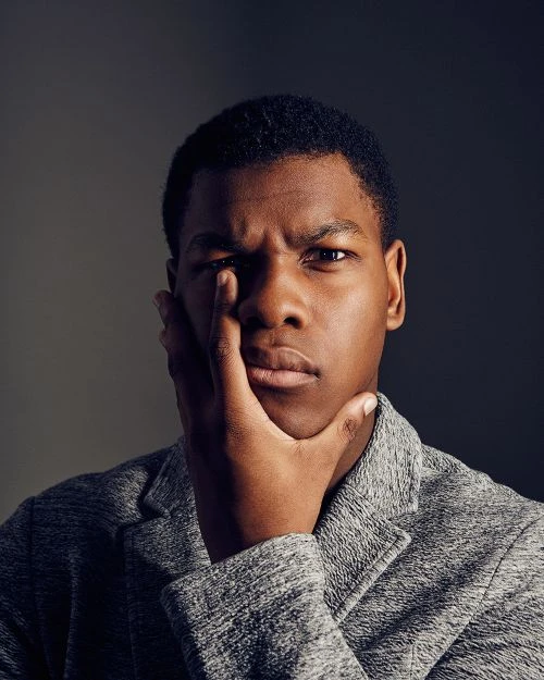 Close-up portrait of a dark-skinned adult male with hand on face, wearing grey jacket against a dark grey backdrop, with serious expression.