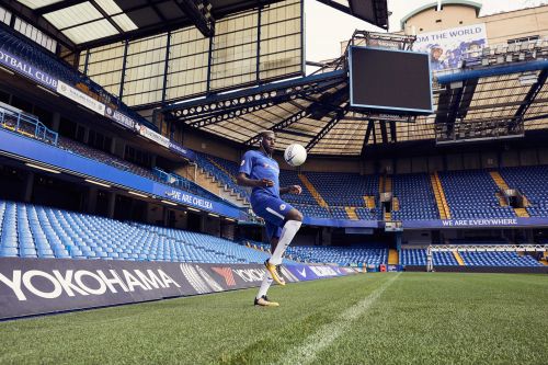 A soccer player wearing a blue uniform kicks a soccer ball in the air on a green field in a stadium filled with blue seats.