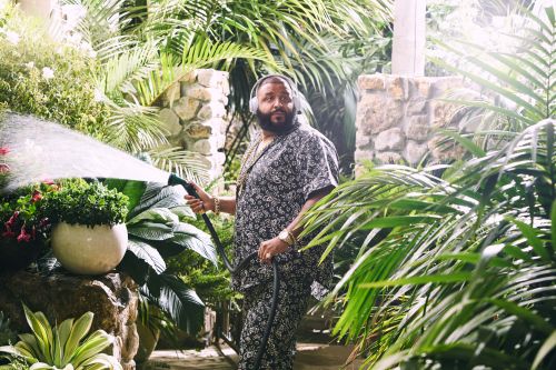 Man waters lush tropical garden. DJ Khaled in patterned shirt and pants, wearing headphones, amidst green foliage, water hose in hand.
