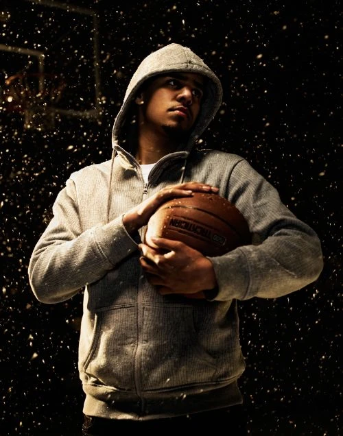 Portrait of adult male in hooded sweatshirt holding basketball in photo studio setting with black backdrop and snow effect.