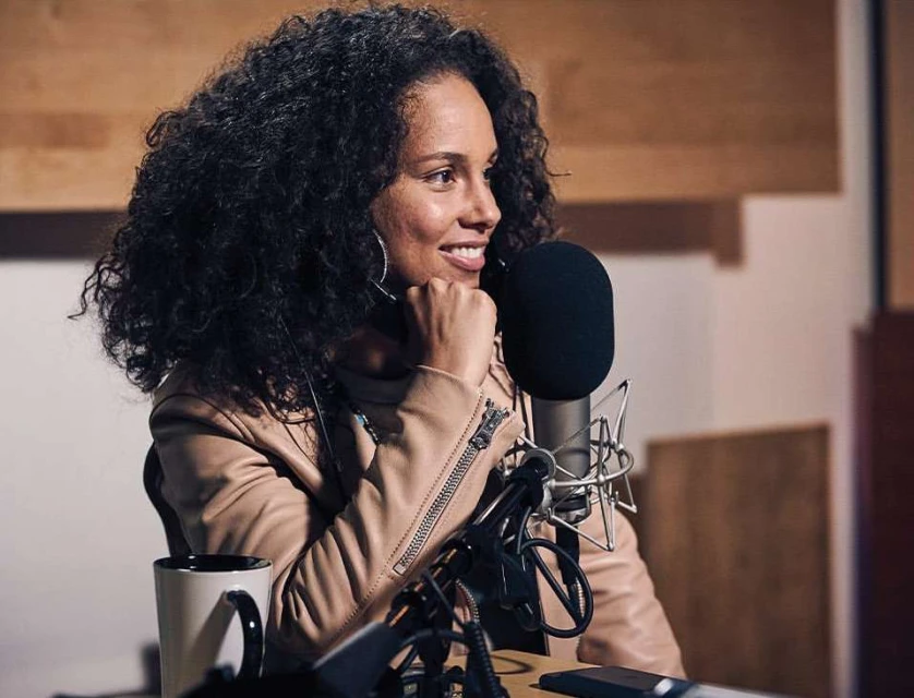 Adult female with curly hair, wearing a jacket, seated at a microphone in a warm-toned recording studio, smiling while working.