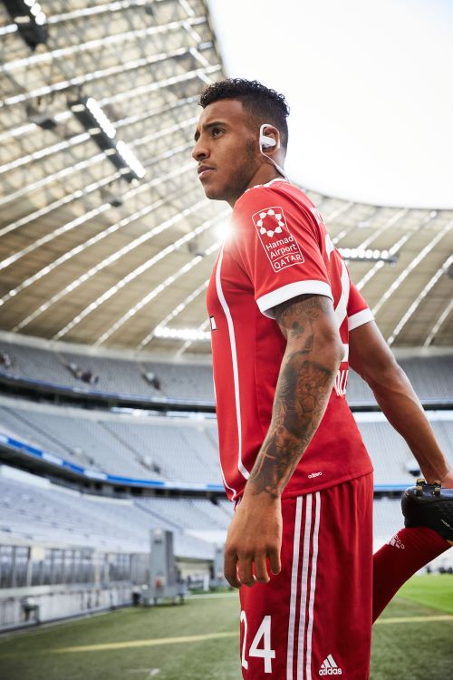Profile of a soccer player in a red and white uniform stretching his leg on a soccer field with the stadium visible in the background.