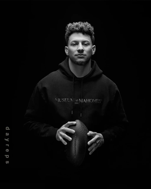 Black-and-white portrait of a man in a hoodie holding an American football, set against a dark background with bright, even lighting.