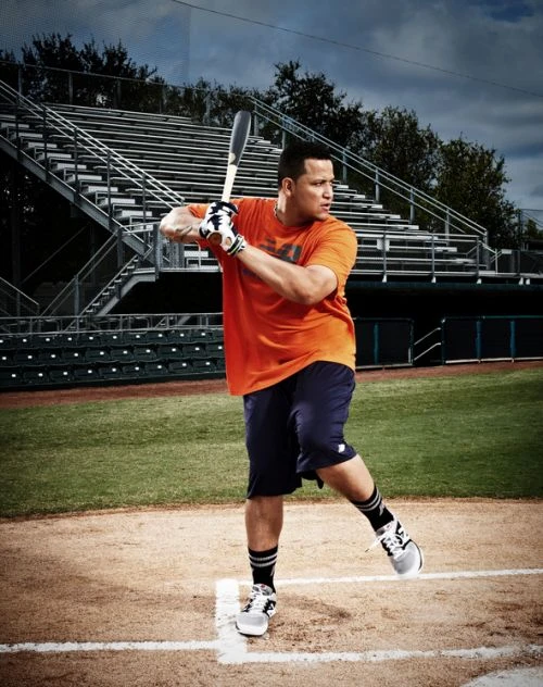 A man in an orange shirt and navy shorts holding a baseball bat on a field, with stadium seating visible in the background, outdoors.