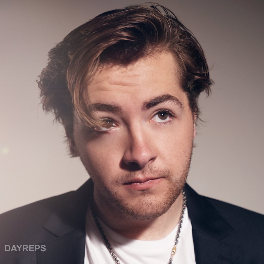 Portrait of a young adult male with brown hair looking upward, wearing a suit jacket and necklace, captured in a photo studio.