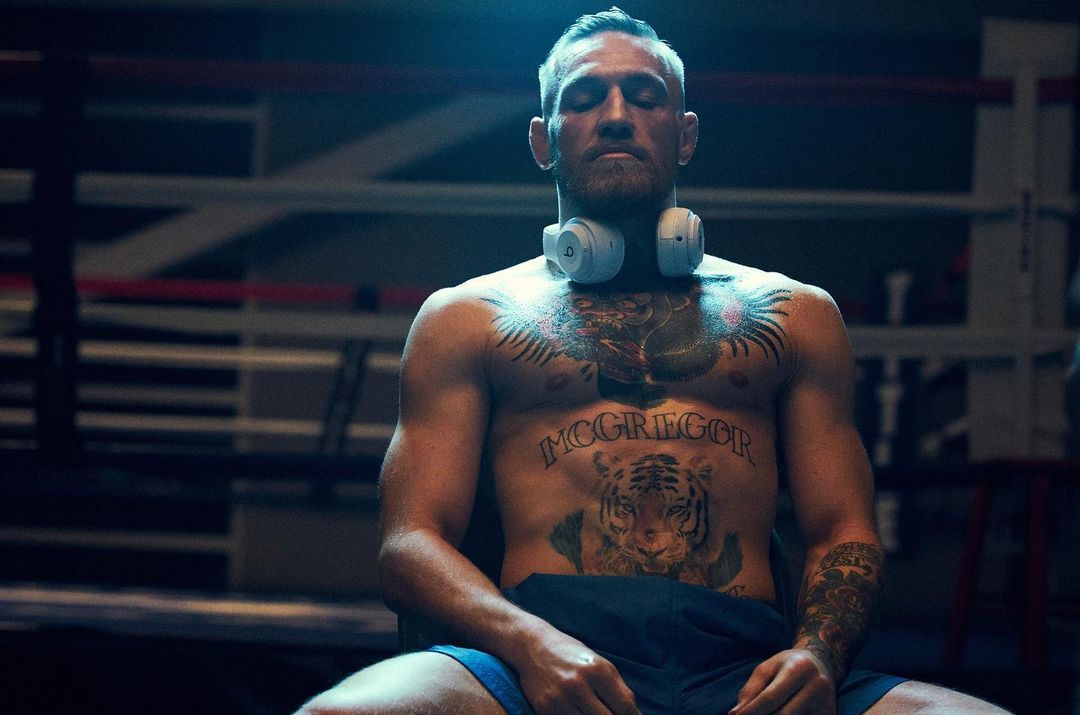 Boxer with tattoos wearing headphones sitting in boxing ring with eyes closed, meditating for a fight or training session.