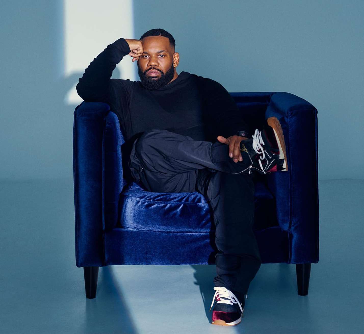 Portrait of a Black man in black clothing, sitting in a dark blue velvet arm chair against a pale blue background in a photo studio.