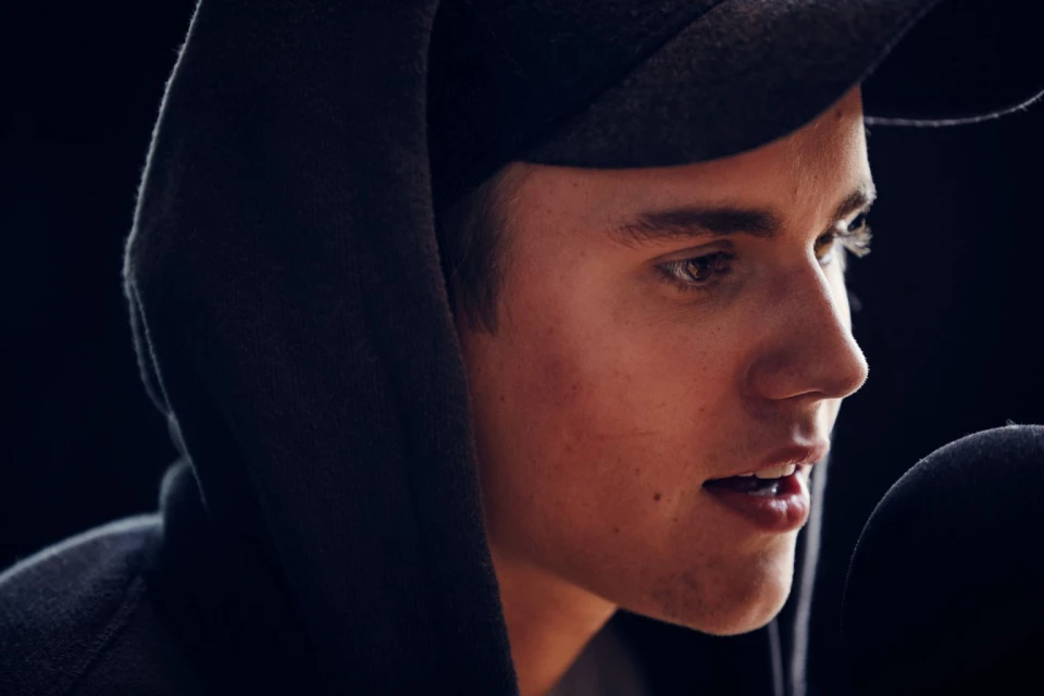 Close-up of a young adult male with a neutral facial expression, wearing a gray hoodie and cap, and a microphone is partially visible.