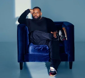 Black man with a beard sitting in a dark blue velvet armchair against a pale blue studio background