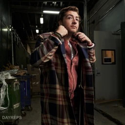 Man wearing a plaid coat adjusting his collar in an industrial hallway with dumpsters in the background.