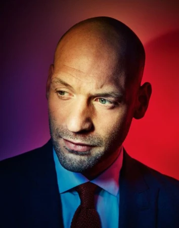 Portrait of a bald man with light eyes wearing a dark suit, light blue shirt, and red tie under colorful red and purple lighting.
