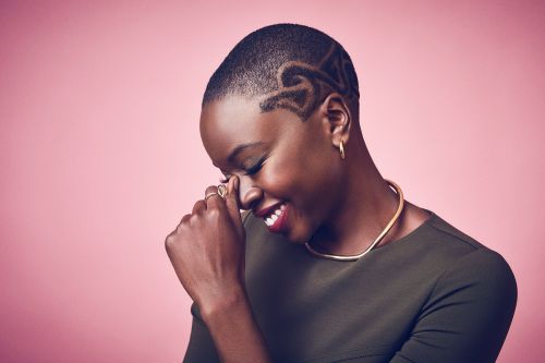 Close-up shot of a smiling Black woman with a decorative buzzcut, gold jewelry, and olive green shirt, against a pink background.