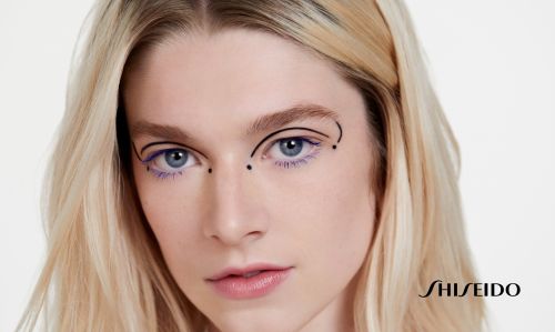 Close-up beauty shot of a blonde woman with a graphic eye makeup design using black lines and purple mascara, against a white backdrop.