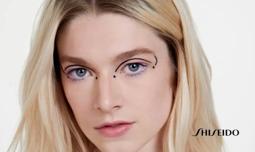 Close-up beauty shot of a blonde woman with a graphic eye makeup design using black lines and purple mascara, against a white backdrop.