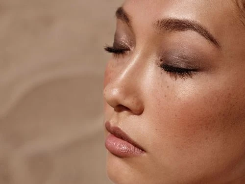 Close-up profile of a woman with closed eyes, natural makeup, and light freckles under warm sunlight.