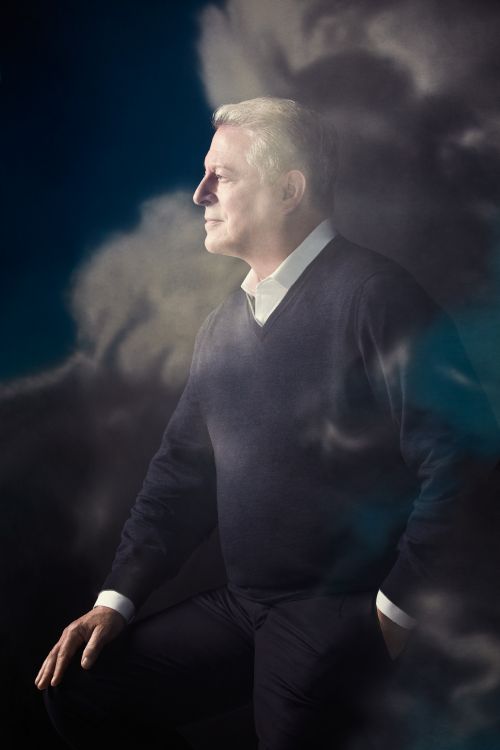 Portrait of a mature man with gray hair, wearing a dark sweater and trousers, posing in front of a cloudy backdrop with soft, diffused lighting.