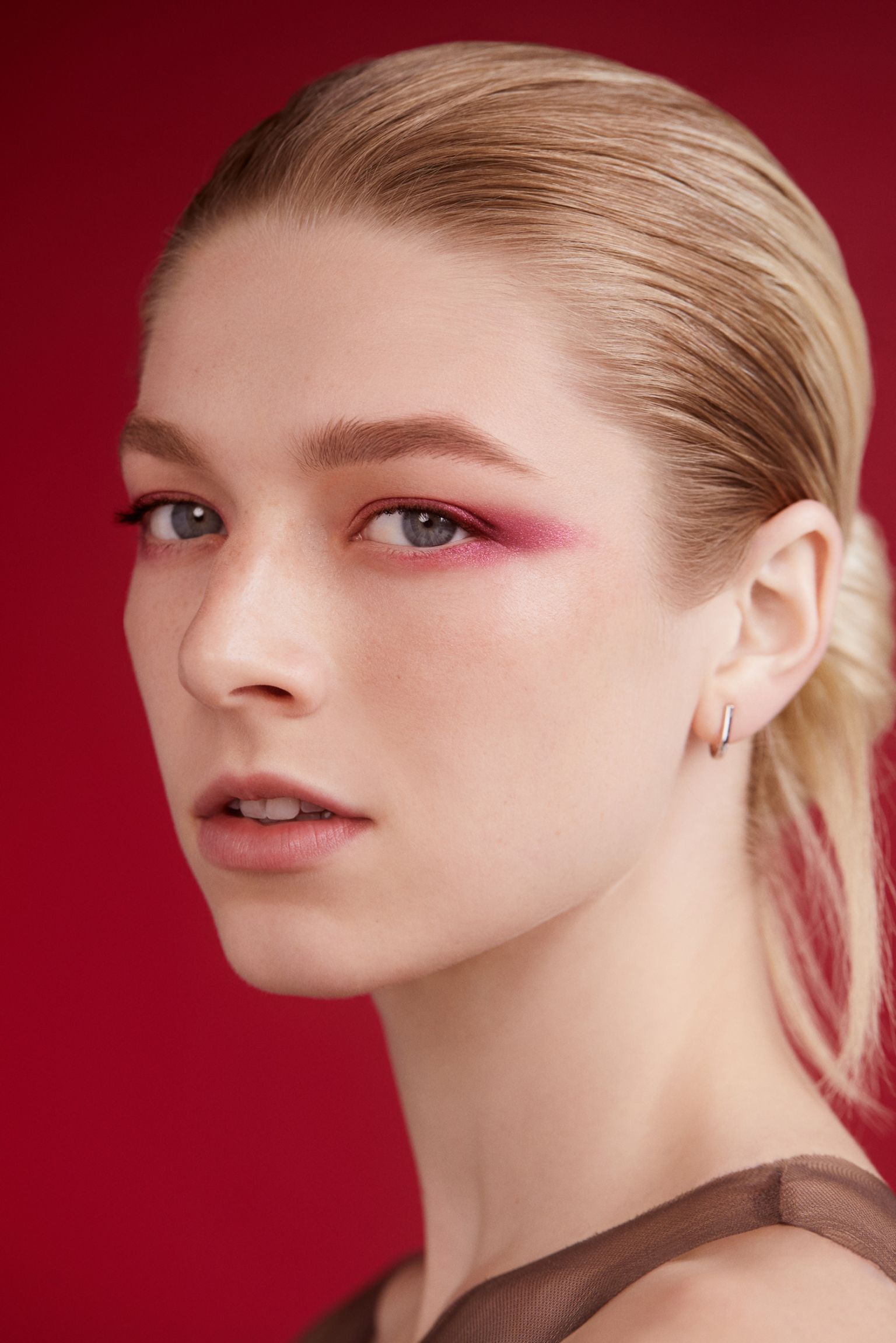 Close-up portrait of a woman with slicked-back blonde hair and pink eye makeup against a solid red background.
