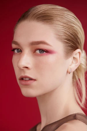 Close-up portrait of a woman with slicked-back blonde hair and pink eye makeup against a solid red background.