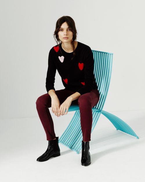 A woman seated on a blue chair, wearing a black sweater with red and pink hearts, maroon trousers, and black ankle boots against a white backdrop.