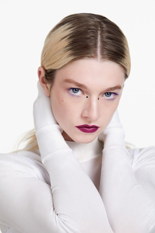 Blonde woman with dark lipstick poses with white gloves and top in a beauty shot against a clean white studio background. Focus on face.