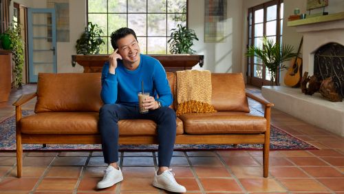 A man with a drink is sitting on a leather sofa in a sunny living room with plants and tile floor, wearing a blue top and white shoes.