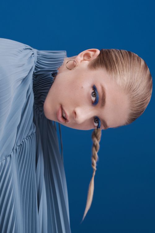Portrait of a young woman with blonde hair in a braid, wearing a pleated blue top, looking at the camera against a blue backdrop.