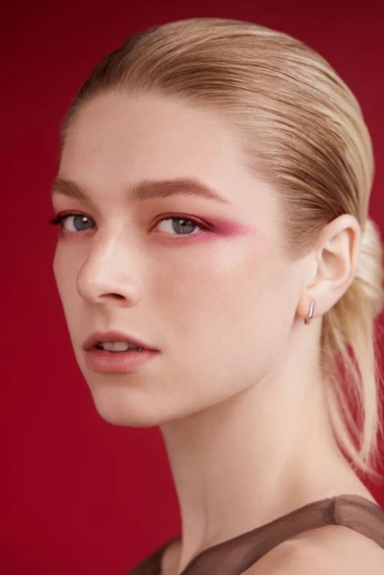 Close-up portrait of a woman with blonde hair and pink eye makeup against a deep red background, showcasing beauty and fashion.