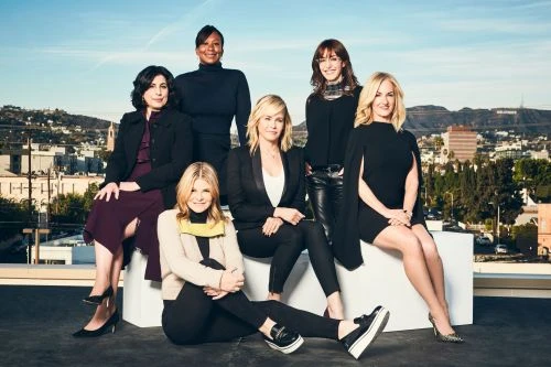 Group portrait of seven adult women posing together on a rooftop with an urban city view in the background on a sunny day.