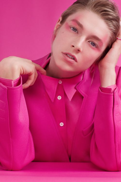 Blonde woman in a pink jacket poses against a pink background, wearing pink makeup around her eyes and cheeks in a studio shot.