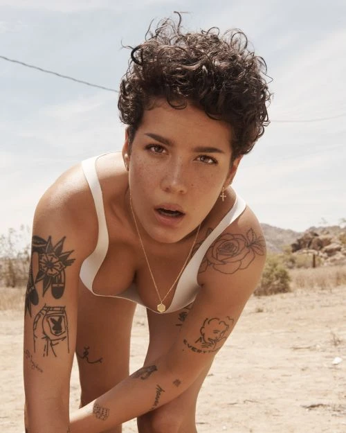 Woman with short curly hair poses outdoors in a white top and gold jewelry, showcasing arm tattoos against a desert background.