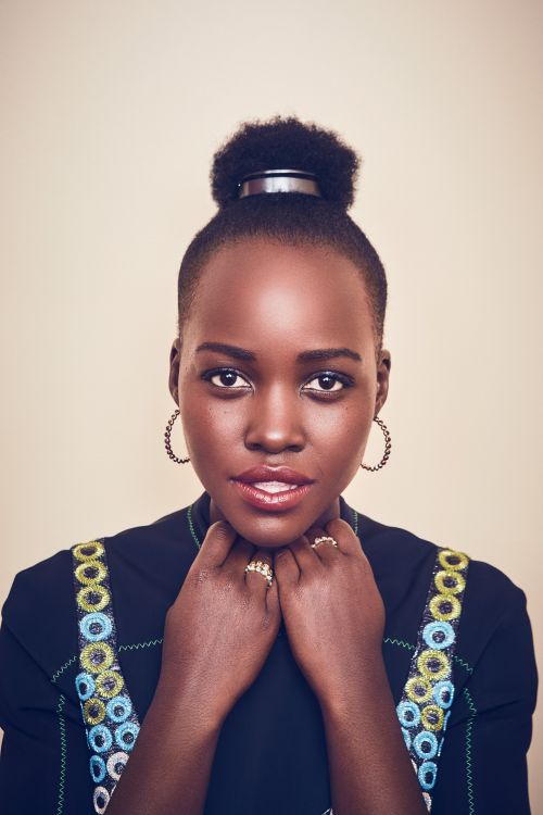 Close-up portrait of an adult Black woman with an updo hairstyle, gold hoop earrings, and a dark-colored, embroidered dress in a studio.