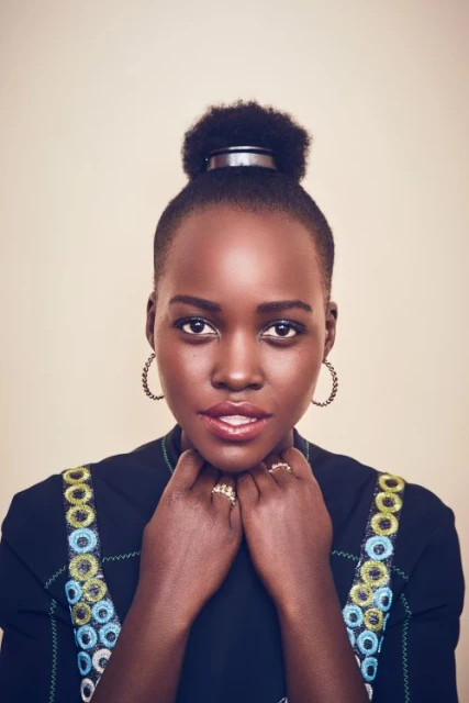 Close-up portrait of an adult Black woman with an updo hairstyle, gold hoop earrings, and a dark-colored, embroidered dress in a studio.