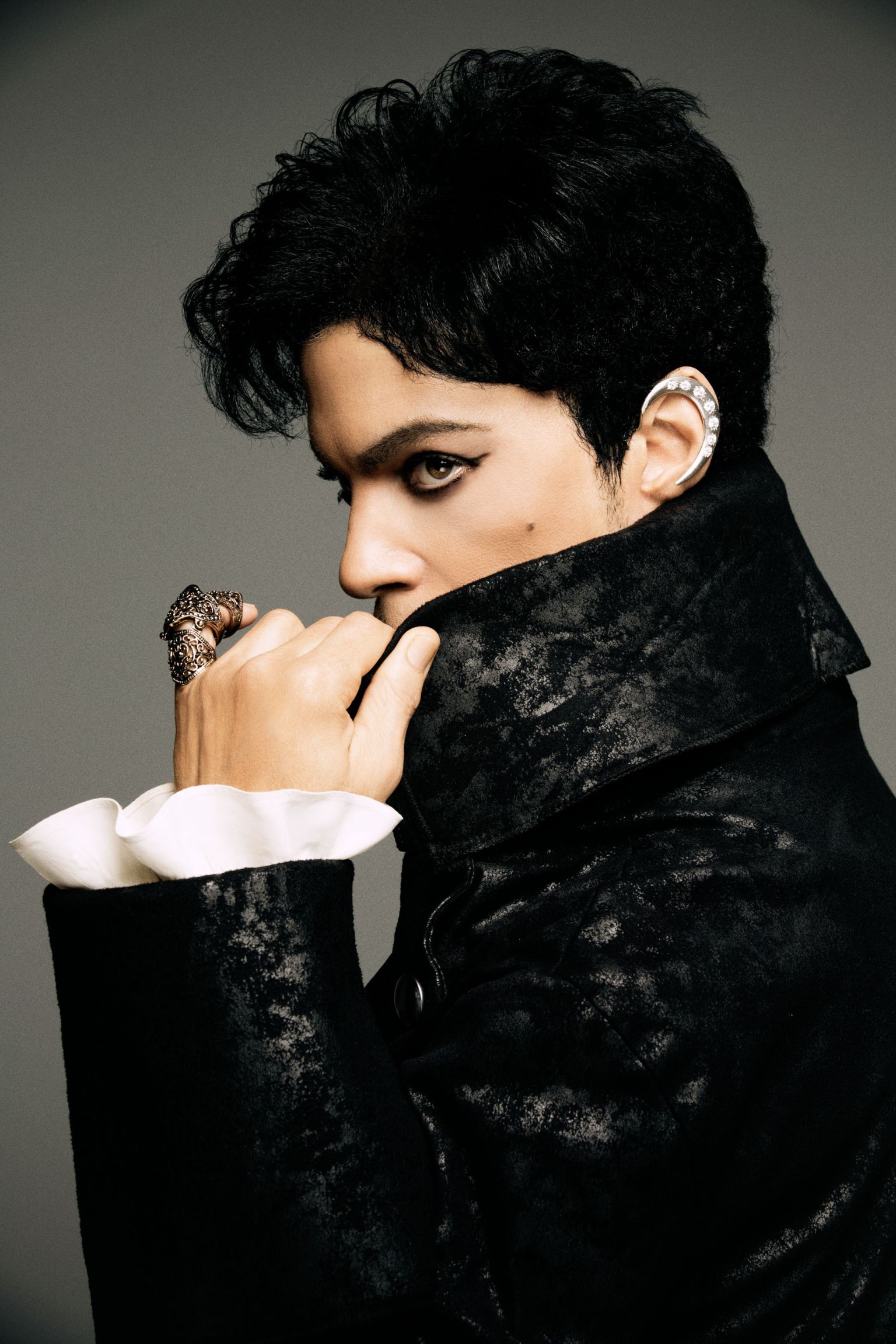 Portrait of a person with dark hair and eyeliner, wearing a black velvet jacket with ruffled white sleeves against a neutral backdrop.
