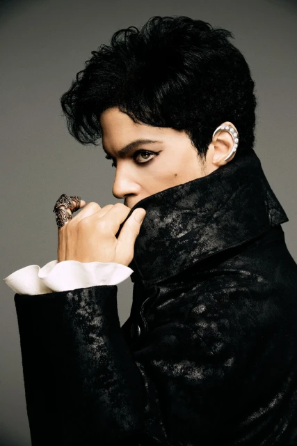 Portrait of a person with dark hair and eyeliner, wearing a black velvet jacket with ruffled white sleeves against a neutral backdrop.