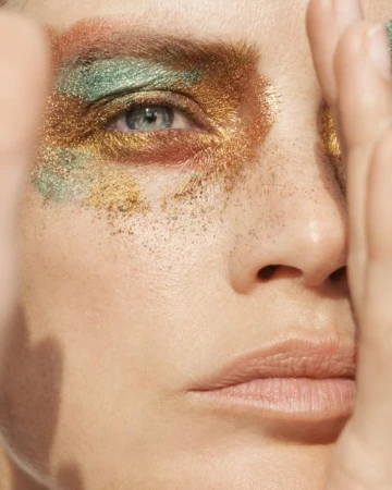 Close-up portrait of a woman with metallic gold, green, and bronze glitter eyeshadow and a hand framing her face.