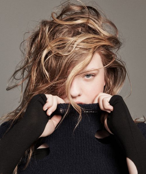 Portrait of a young woman with blonde hair partially covering her face, wearing a dark sweater with cut-out details, against a grey background.