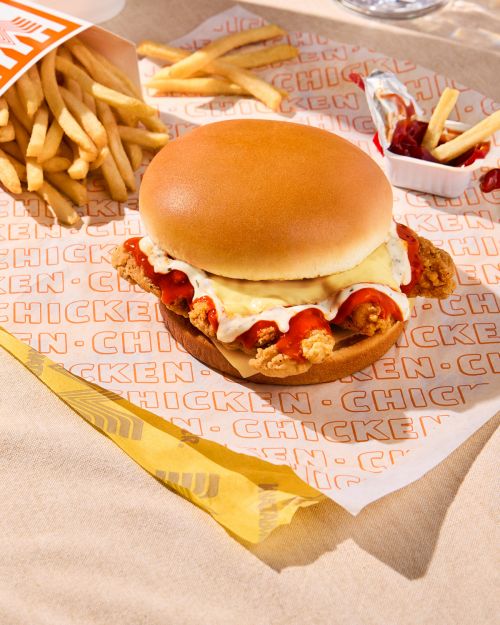 Overhead shot of a chicken sandwich with spicy sauce and cheese, served with fries, fast food packaging, and condiments on a branded paper.