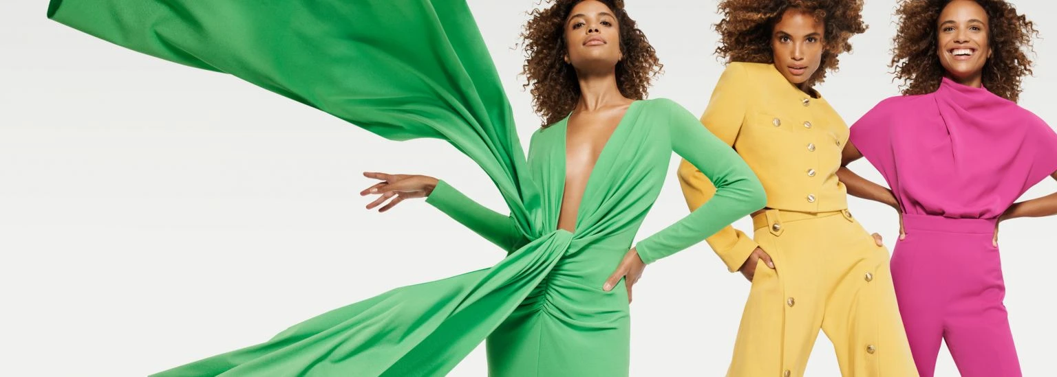 Three women pose in brightly colored, fashionable outfits against a plain white background, creating a vibrant and contemporary fashion image.
