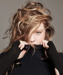 Close-up portrait of a woman with tousled blonde-streaked hair pulling up the collar of a black turtleneck sweater over her lower face.