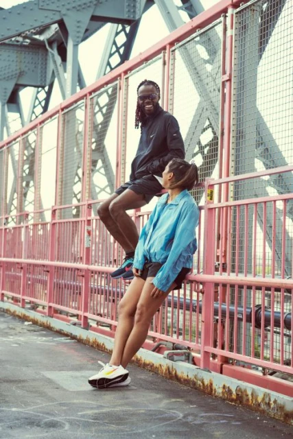 An athletic couple, a man and a woman, are posing on a pink bridge. The man is sitting on the railing and the woman is leaning on it.