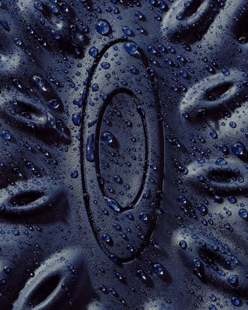 Close-up of a textured navy-blue surface with an embossed oval logo covered in water droplets, creating a wet and abstract design.