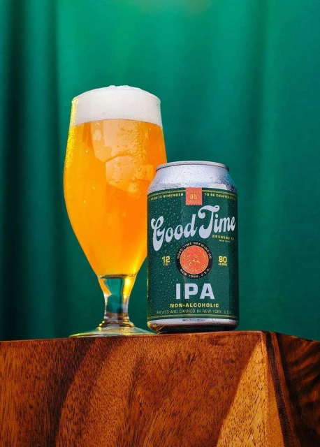 A non-alcoholic IPA beer is positioned next to a beer glass filled with a golden brew, set against a backdrop of rich green fabric.